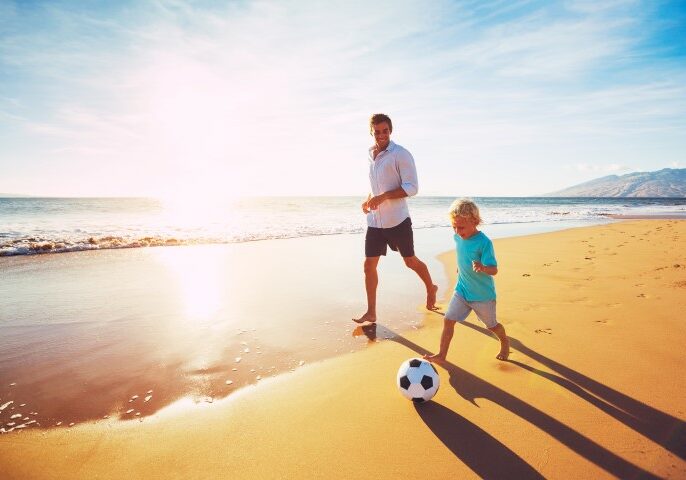 A dad playing soccer with his son on the beach